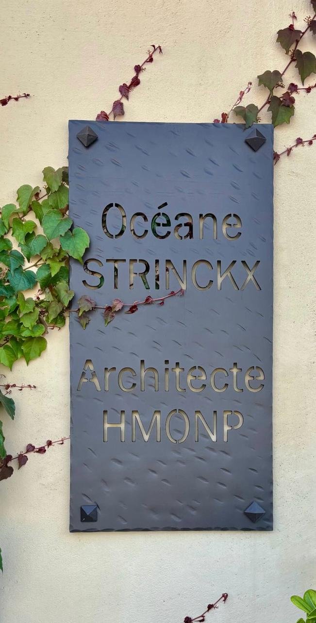 Plaque en métal de l'agence : Océane Strinckx, Architecte HMONP, sur un mur ensoleillé couvert de lierre